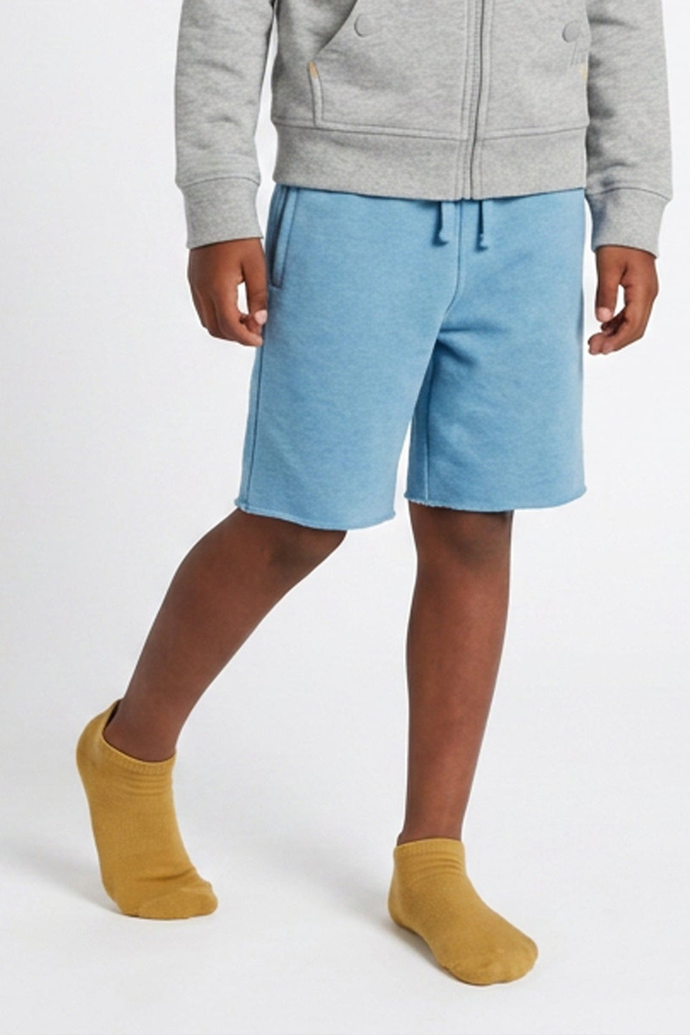 A child wearing light blue sweat shorts, a gray zip-up hoodie, and mustard yellow socks, standing against a plain white studio background.