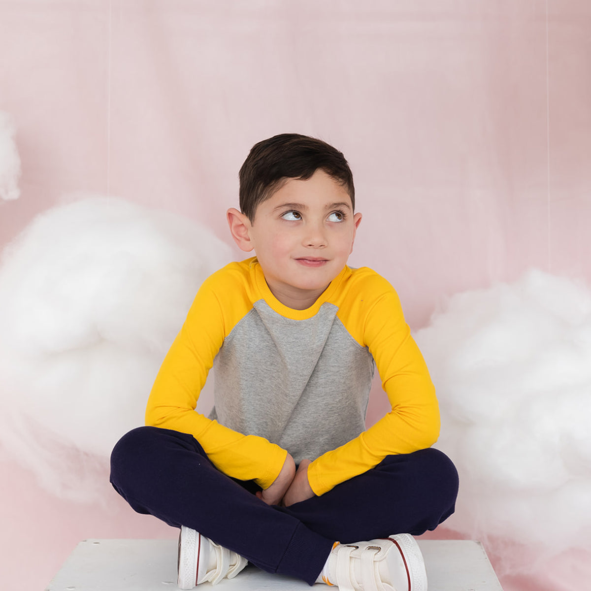 A boy sitting crossed legged on the floor wearing a gray and yellow long sleeve baseball tee