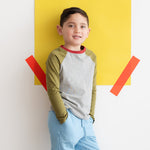 A young boy wearing a gray and green long sleeve baseball tee leaning against a colorful wall