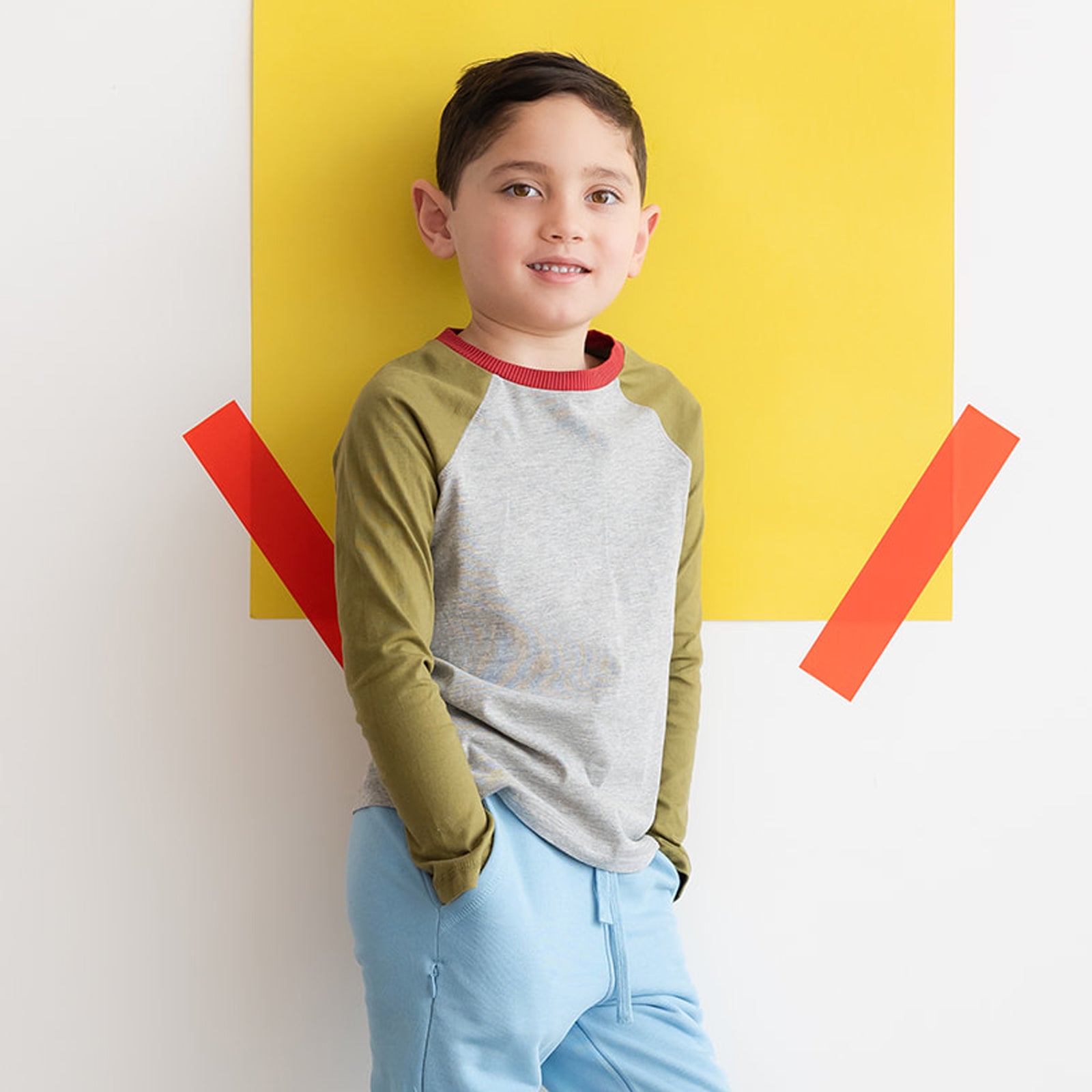 A young boy wearing a gray and green long sleeve baseball tee leaning against a colorful wall