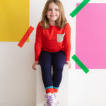 a young girl wearing a red long sleeve tee with a lightning bolt print pocket sitting on a bench against a colorful wall