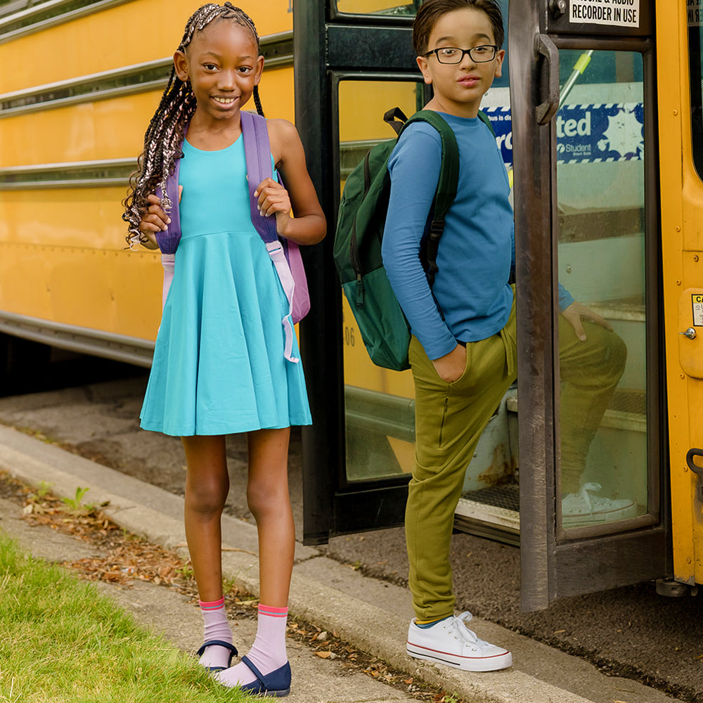 a girl wearing a turquoise dress and a boy wearing a blue shirt and olive green sweatpants getting on a school bus