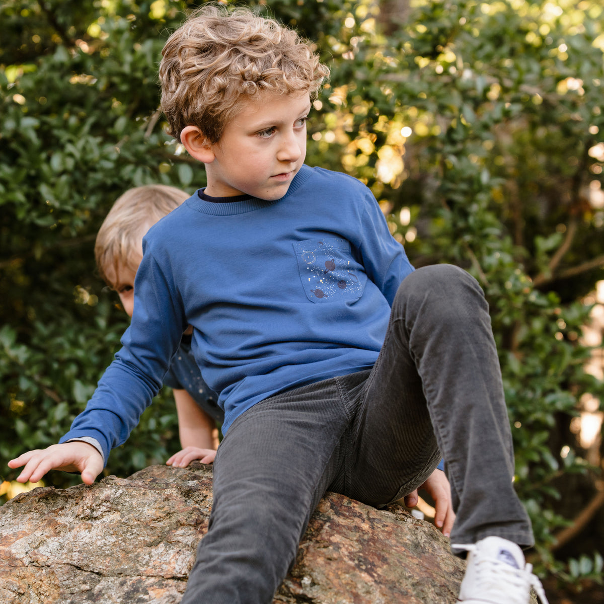 a young boy sitting on a rock wearing a blue long sleeve tee with a galaxy print on the pocket
