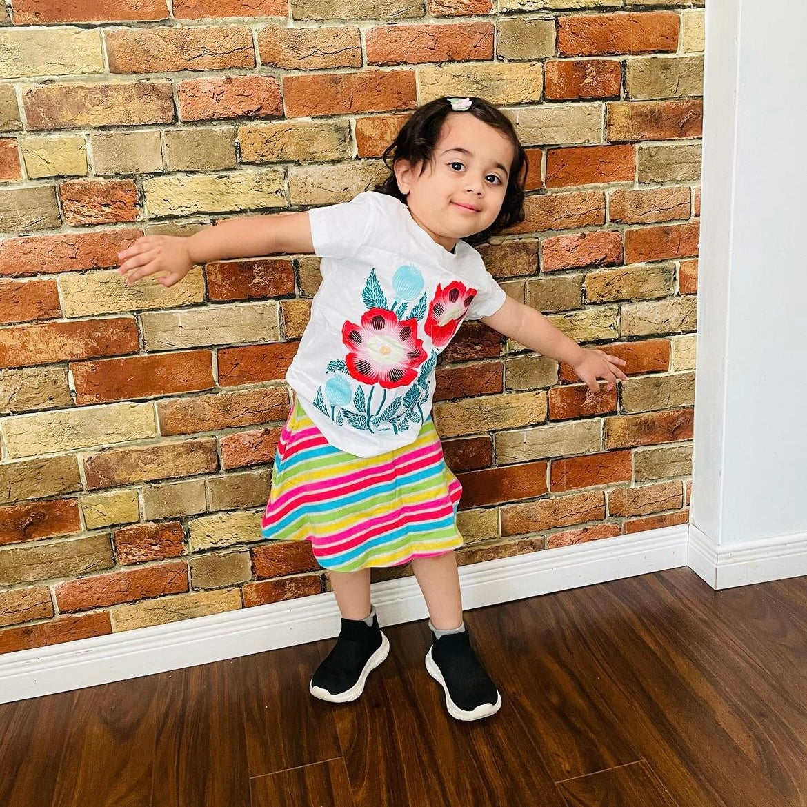 A young child wear a white shirt with a floral print and striped skirt against a brick wall