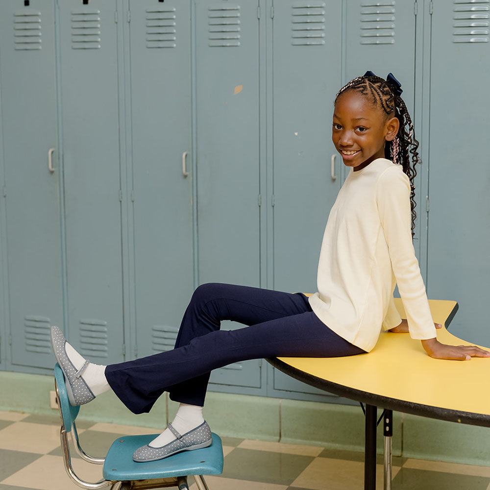 Girl wearing a long sleeve ivory shirt with navy blue flared leggings sitting on a desk in front of lockers